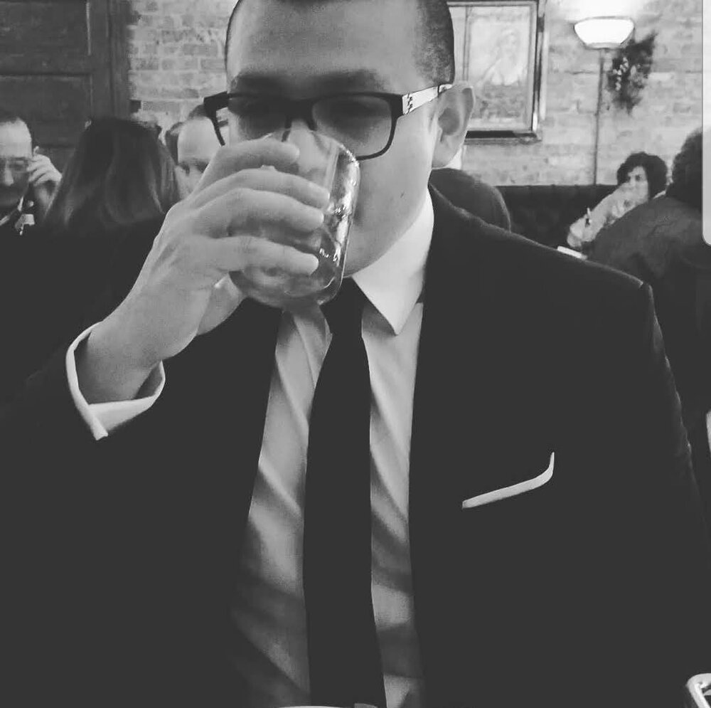 A black and white photo of a Latino man with glasses, drinking from a highball glass.